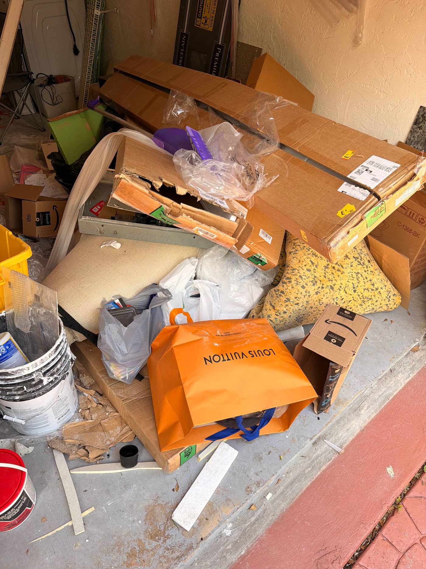 A garage doorway blocked by a collapsed pile of flattened boxes, packing material, bags and offcuts of timber spilling onto the driveway pavers