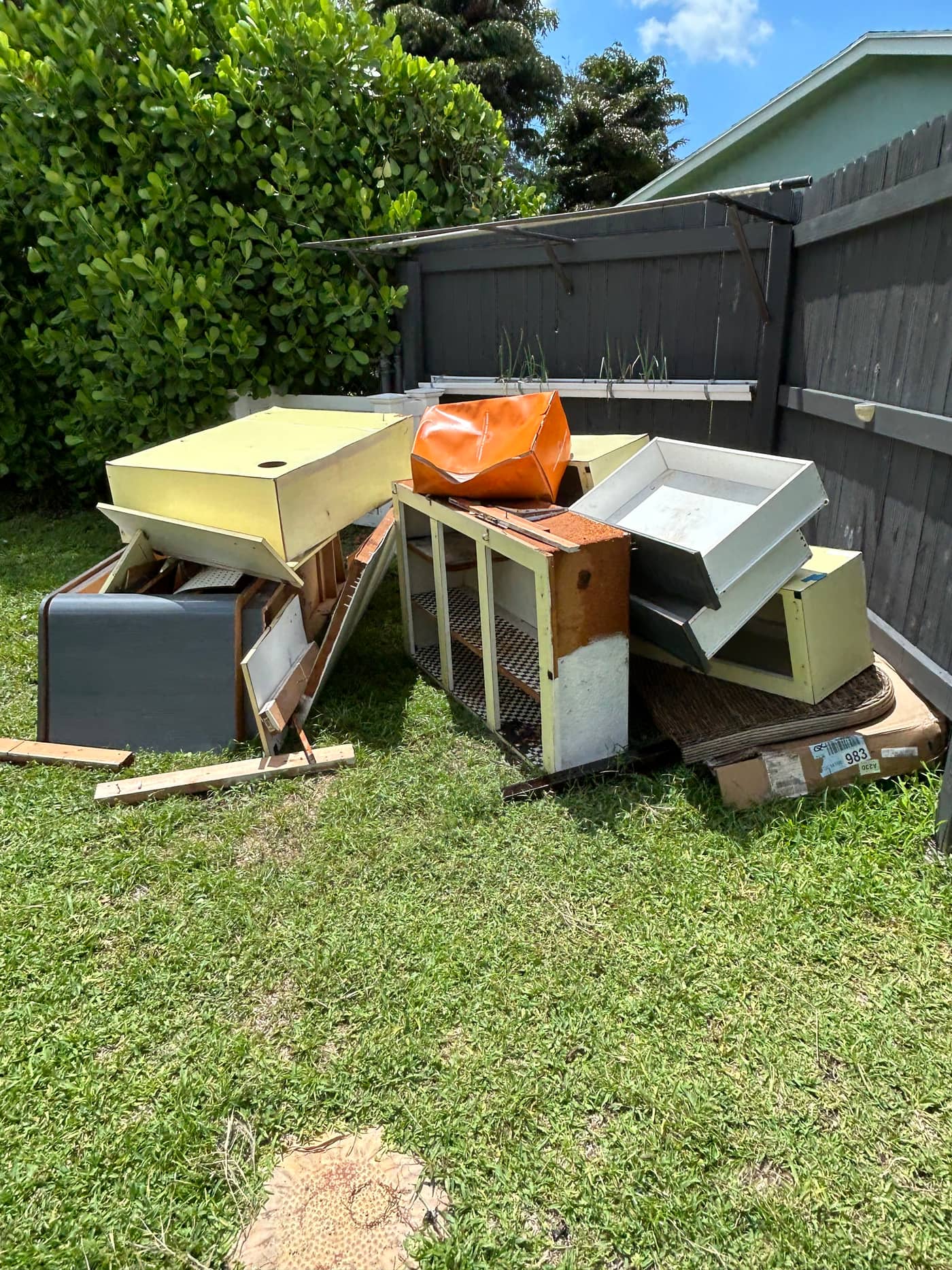 Old kitchen cabinets, a countertop, a filing cabinet and a bagged item stacked on the grass against a fence